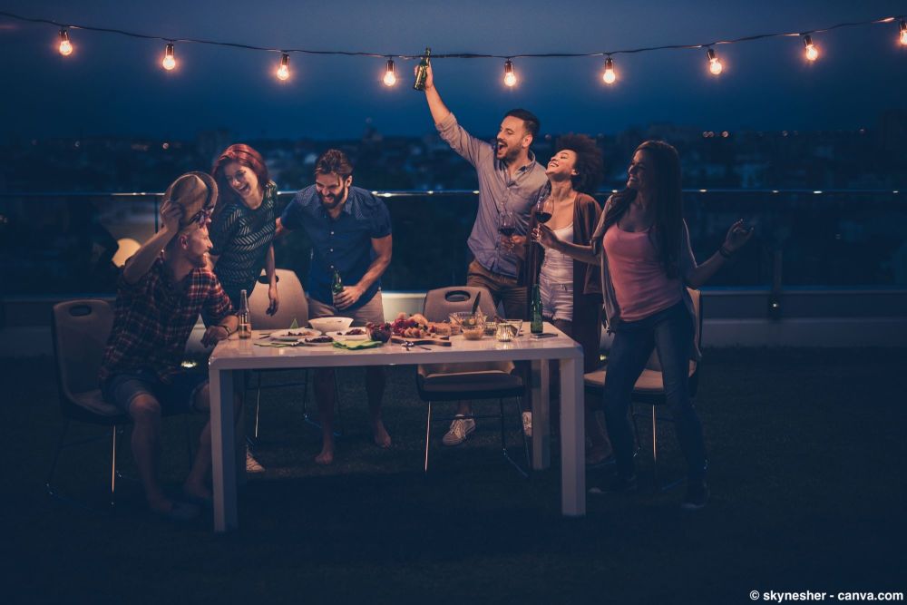 Freunde feiern abends auf einer Dachterrasse mit Lichterkette, sitzen und tanzen um einen gedeckten Tisch.