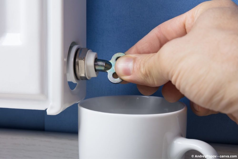 Hand entlüftet einen Heizkörper mit einem Schlüssel, darunter steht eine Tasse zum Auffangen von Wasser.