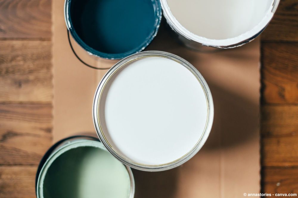 Geöffnete Farbdosen mit weisser, blauer und grüner Wandfarbe auf einer abgedeckten Holzoberfläche.