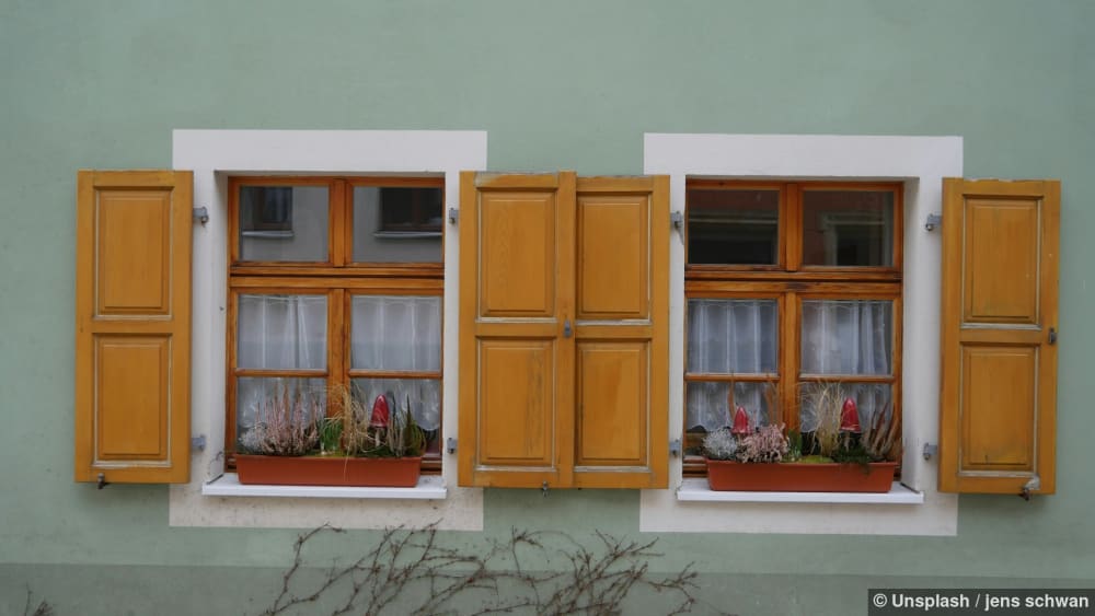 Zwei Fenster mit hölzernen Fensterläden und Pflanzen sind zu sehen