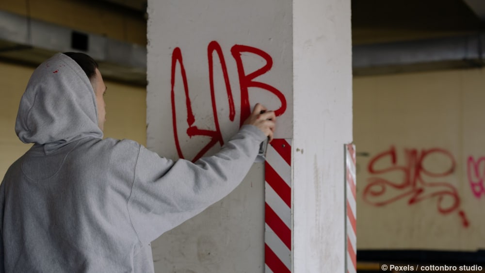 Kapuzentragender Künstler sprüht Graffiti an eine Betonwand in städtischer Umgebung