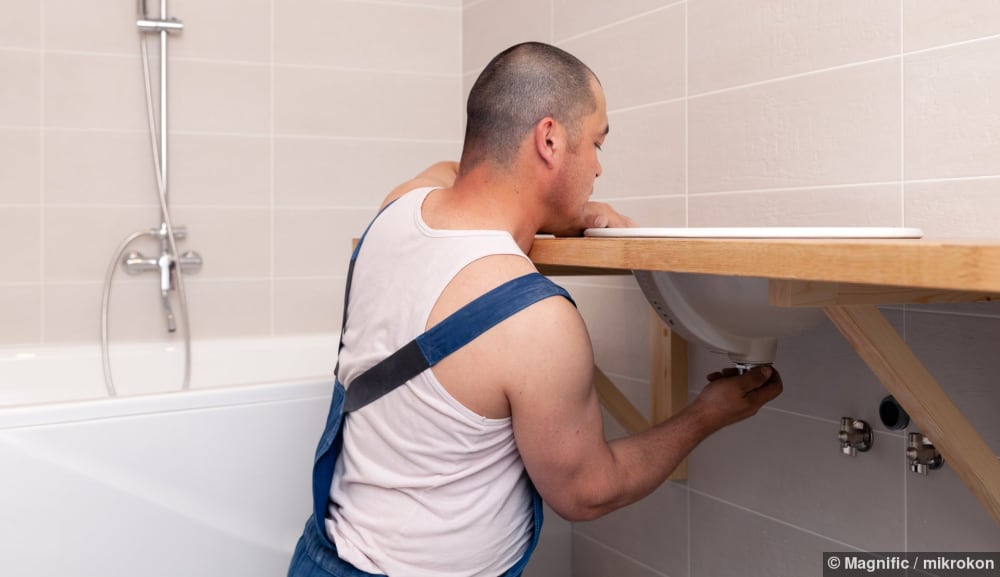 Sanitärinstallateur in blauer Latzhose repariert ein Lavabo in einem Badezimmer mit gefliester Wand