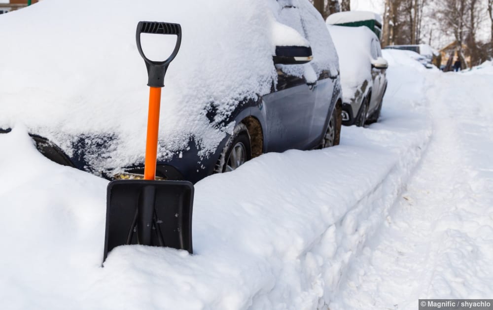 Eine Schneeschaufel steht vor einem eingeschneiten Auto